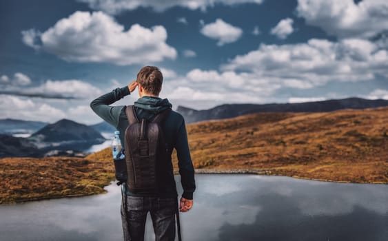 Young adult man hiking looking at view with lake and mountaing range in background, wearing a backpack, rear view, standing, volda norway