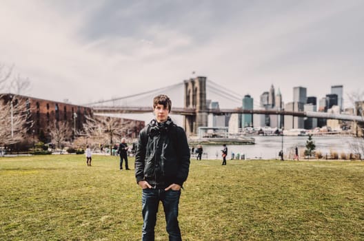 Young adult man standing in brooklyn on a meadow looking at camera, brooklyn bridge and skyline of manhattan cityscape new york in background, nyc, america