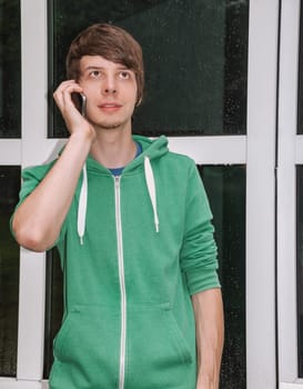 young man with brown hair and a beard is talking on the phone with his smartphone in front of his door with raindrops, wearing a green sweater, vertical
