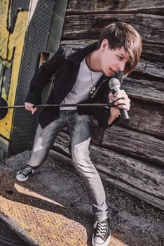 Young adult singer, with brown long hair and beard performing, rock musician with microphone outdoors in front of a wooden wall, standing, looking away, wearing jeans and t-shirt