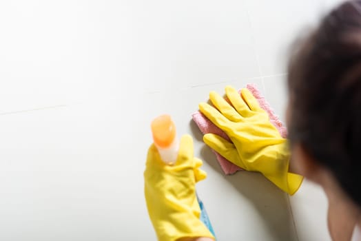 Asian woman wearing yellow rubber glover with cloth rag and detergent spray cleaning floor at home in living room, Female hands wash cleaner, housework and housekeeping concept