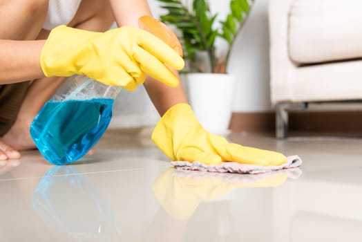 Asian woman wearing yellow rubber glover with cloth rag and detergent spray cleaning floor at home in living room, Female hands wash cleaner, housework and housekeeping concept