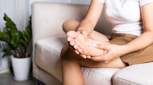 Foot pain, Asian woman sitting on sofa feeling pain in her foot at home, female suffering from feet ache use hand massage relax muscle from soles in home interior, Healthcare and podiatry medical