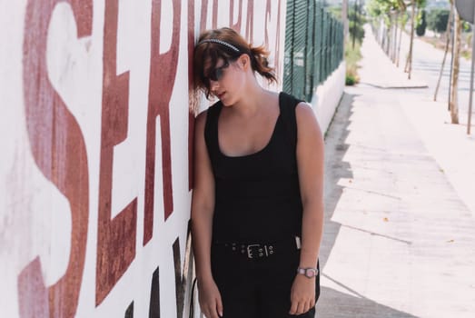 Young woman with black orange hair, wearing sunglasses and black casual clothing, leaning exhausted against wall to take a break from a long hike, front view, horizontal