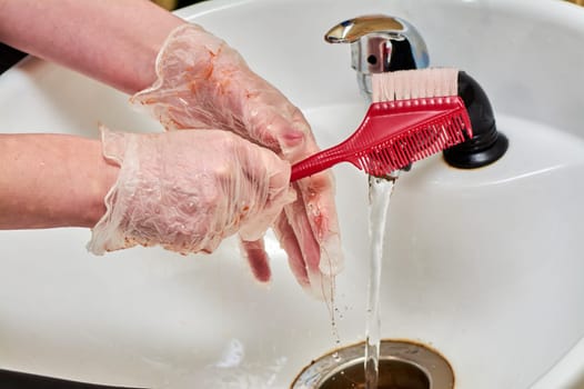 Women's hands in hair coloring gloves wash a comb under a stream of water in a white sink. High quality photo