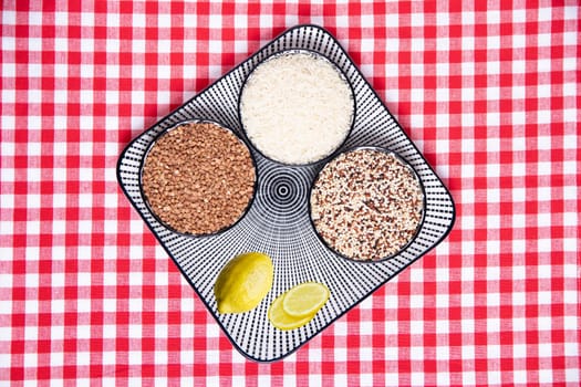 Vegetable food. Close-up of a decorative plate with three bowls with buckwheat, rice, quinoa and lemon on a red checkered tablecloth. Top view with space for design.