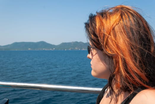 Woman with black orange hair, wearing sunglasses sitting on a ship looking at the mediterranean sea, island in the background, close-up horizontal