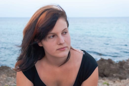 Young woman with black orange hair sitting in front of the mediterranean sea, looking away, close-up, horizontal