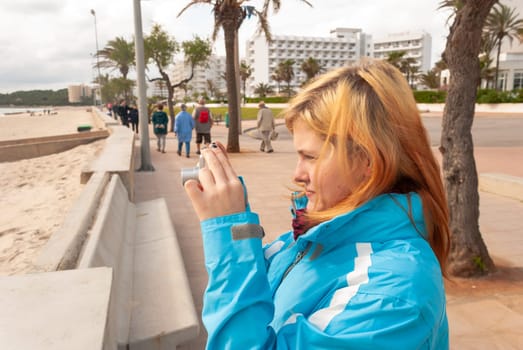 Young woman with blond hair, wearing a blue jacket standing at the promenade, taking photos of the beach in Cala Millor, Majorca