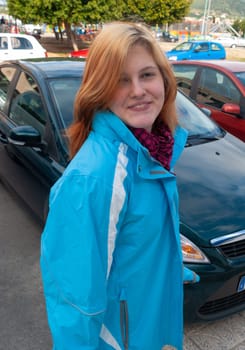 Young woman with orange blond hair, wearing a blue jacket smiling, looking at camera, standing on a sidewalk in front of cars at a car rental in majorca, spain
