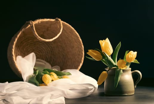 Still life with bouquet of yellow tulip flowers in old vintage jug, in beam of light on dark background.Art photography.