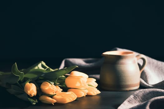 Still life with bouquet of yellow tulip flowers in old vintage jug, in beam of light on dark background.Art photography.