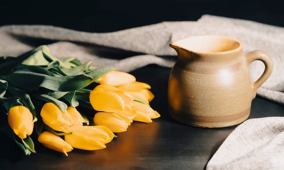 Still life with bouquet of yellow tulip flowers in old vintage jug, in beam of light on dark background.Art photography.