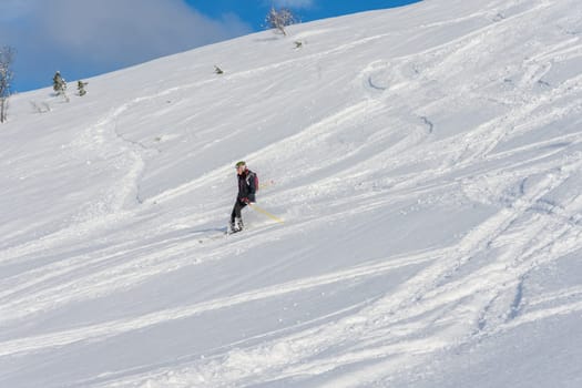 Woman with brown hair, wearing ski wear and a bagpack skiing down a beautiful snow hill with plenty of tracks on the snow, view from the side from far away, stryn norway, horizontal