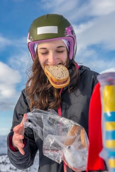 Woman with brown hair, wearing ski wear, taking a break from skiing in Stryn, Norway, Holding a bag and biting in a bread with eyes closed, vertical