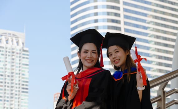 Two young women celebrating their graduation, holding academic degree and standing outdoors in the city