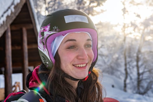 Woman with brown hair, wearing ski helmet and ski wear smiling while looking at view from top of the mountain before skiing, early in the morning with sunlight and lens flare, view from the side, horizontal
