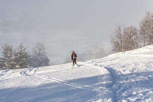 Woman with brown hair, wearing black ski wear and helmet skiing down the top of a mountain, rear view, beautiful snow landscape with trees, valley in the distance, fog, horizontal