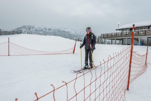 Skiing woman with brown hair smiling, wearing black ski wear and a helment, entering ski lift area early in the morning, fence in forefront, building in background, side view, horizontal