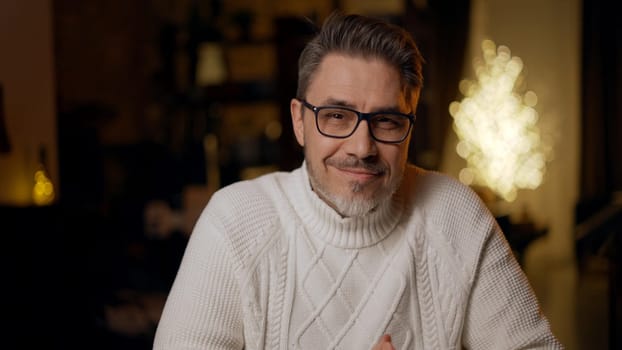 Portrait of mature age, middle age, mid adult casual man in 50s, confident happy smiling. Dark cosy room at home, winter, Christmas.