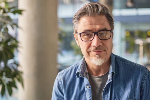 Portrait of happy middle aged man in glasses, standing in office lobby wearing casual shirt.