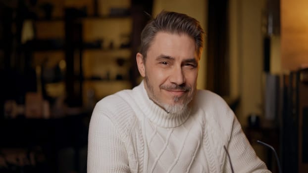 Portrait of mature age, middle age, mid adult casual man in 50s, confident happy smiling. Dark cosy room at home, winter