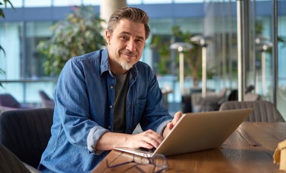 Happy middle age businessman using laptop computer in office. Older casual man working online on terrace of cafe, smiling.