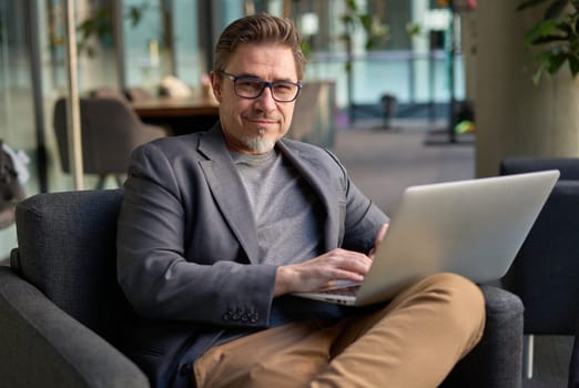 Businessman using laptop computer in office lobby or on cafe terrace. Happy middle aged man, entrepreneur, small business owner working online.