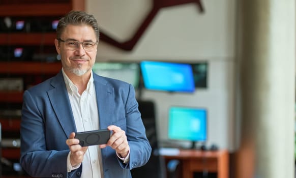 Business portrait - confident businessman standing in office, using phone. Happy mid adult man in shirt and jacket, smiling. Bearded, gray hair.