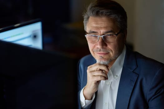 Businessman working with computer at office desk, thinking, smiling. Portrait of happy mid adult man in shirt and jacket, smiling. Bearded, glasses.