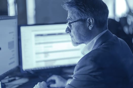 Businessman working with computer at office desk, thinking. Portrait of mid adult man in shirt and jacket, smiling. Bearded, glasses.
