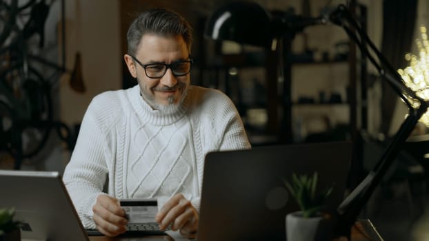 Happy mid adult man at home shopping on the internet using laptop computer paying online with credit card. Winter, Christmas, dark cosy room.