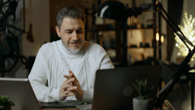 Happy mid adult man working with laptop computer in home office. Winter, Christmas, dark cosy room.