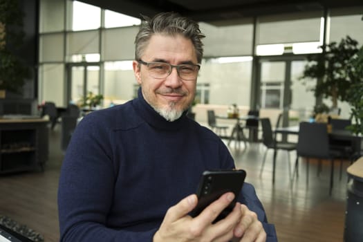 Smiling middle age man using phone sitting in restaurant or cafe.