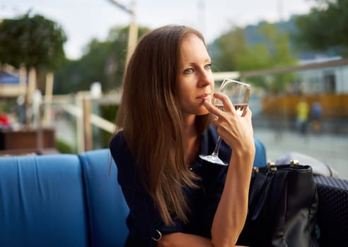 Elegant young woman sitting outdoor in restaurant terrace, smiling drinking wine.