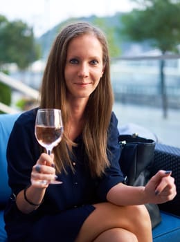 Elegant young woman sitting outdoor in restaurant terrace, smiling drinking wine.