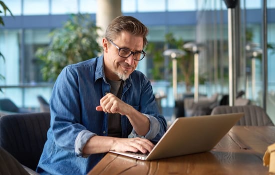 Happy middle age businessman using laptop computer in office. Older casual man working online on terrace of cafe, smiling.