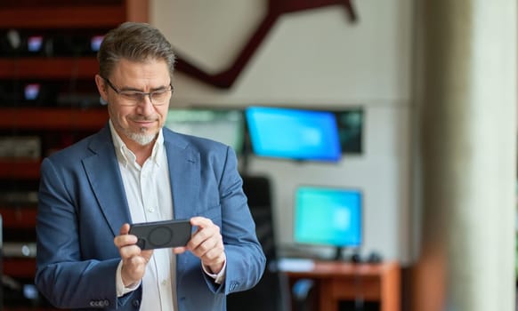 Business portrait - confident businessman standing in office, using phone. Happy mid adult man in shirt and jacket, smiling. Bearded, gray hair.