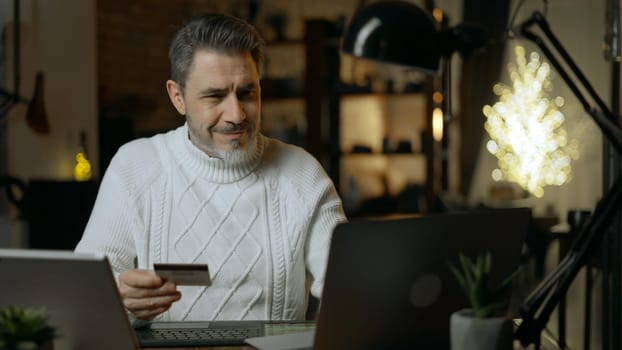 Happy mid adult man at home shopping on the internet using laptop computer paying online with credit card. Winter, Christmas, dark cosy room.