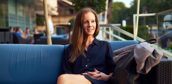 Elegant young woman sitting outdoor in restaurant terrace, smiling.