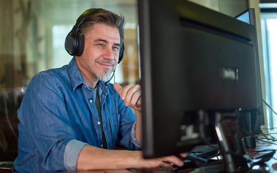 Businessman working with computer in office talking on headset. Man sitting at desk in front of screen, smiling, happy. Customer service support operator