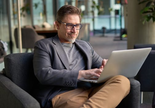 Businessman using laptop computer in office lobby or on cafe terrace. Happy middle aged man, entrepreneur, small business owner working online.