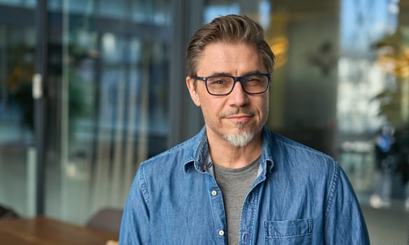 Portrait of happy middle aged man in glasses, standing in office lobby wearing casual shirt.