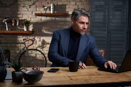 Older businessman in jacket working with laptop computer at desk in trendy loft office.