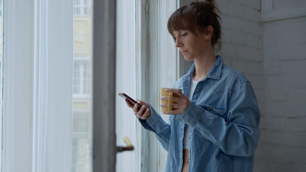 Adult woman standing in window at home daydreaming drinking coffee and checking phone at morning.