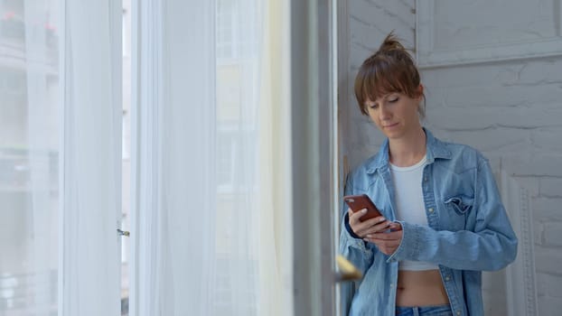 Adult woman standing in window at home daydreaming and checking phone at morning.
