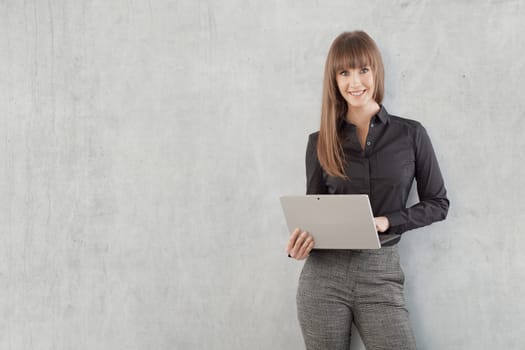 Happy young businesswoman with laptop computer standing against gray concrete wall. Copy space for your text.