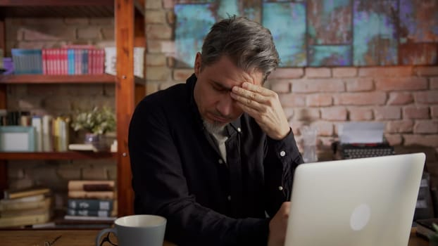 Mature man working alone from home. Sitting at desk teleworking with laptop in home office. Looking tired and troubled, thinking and having problem to solve.