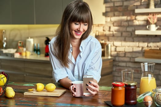 Happy young attractive white woman in man's shirt using mobile phone while having breakfast in kitchen at home at the morning, smiling.