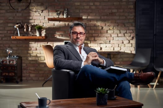 Stay at home - smart looking older man sitting in armchair in home office reading book.